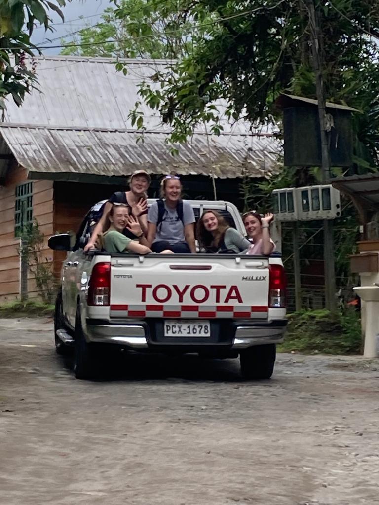 People riding in the bed of a white Toyota pickup truck parked on a dirt street beside a brick building.
