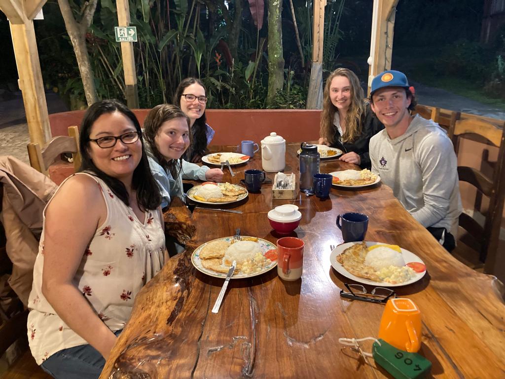 People smiling around a wooden dining table with plates of food in the evening.