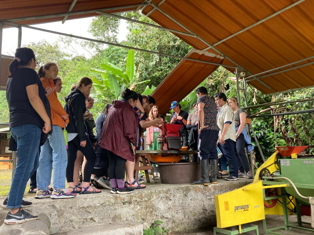 Students standing around coffee-processing equipment under a shelter.