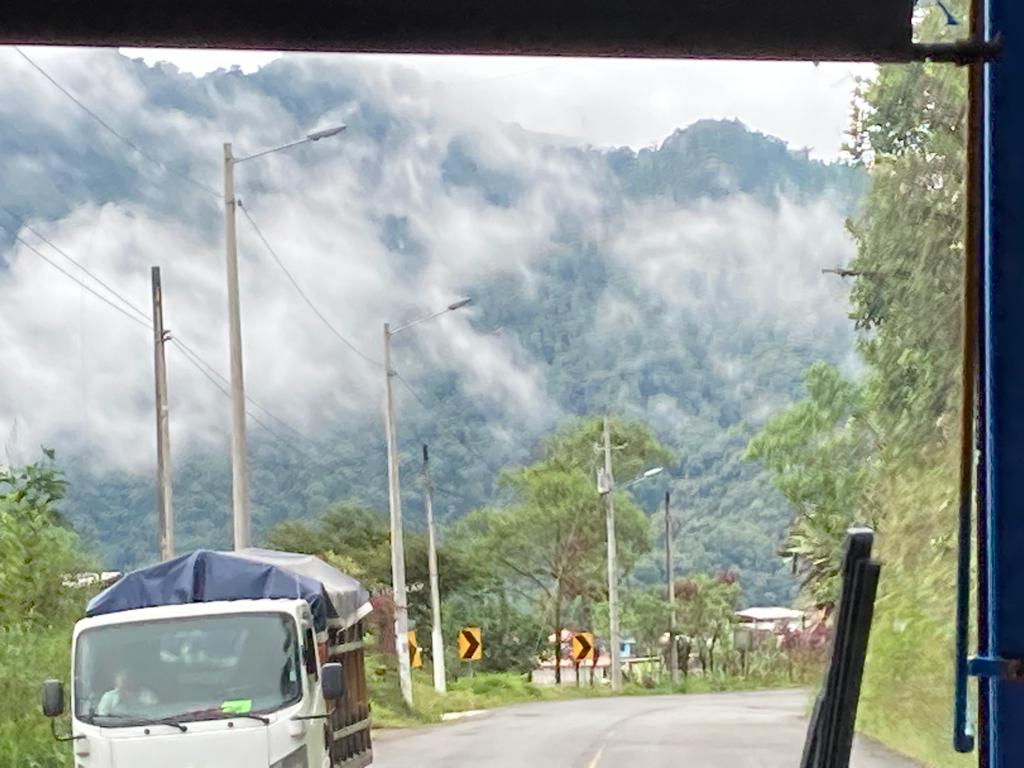 View through a vehicle window of a white truck on a rural road with cloud-covered green mountains in the background.