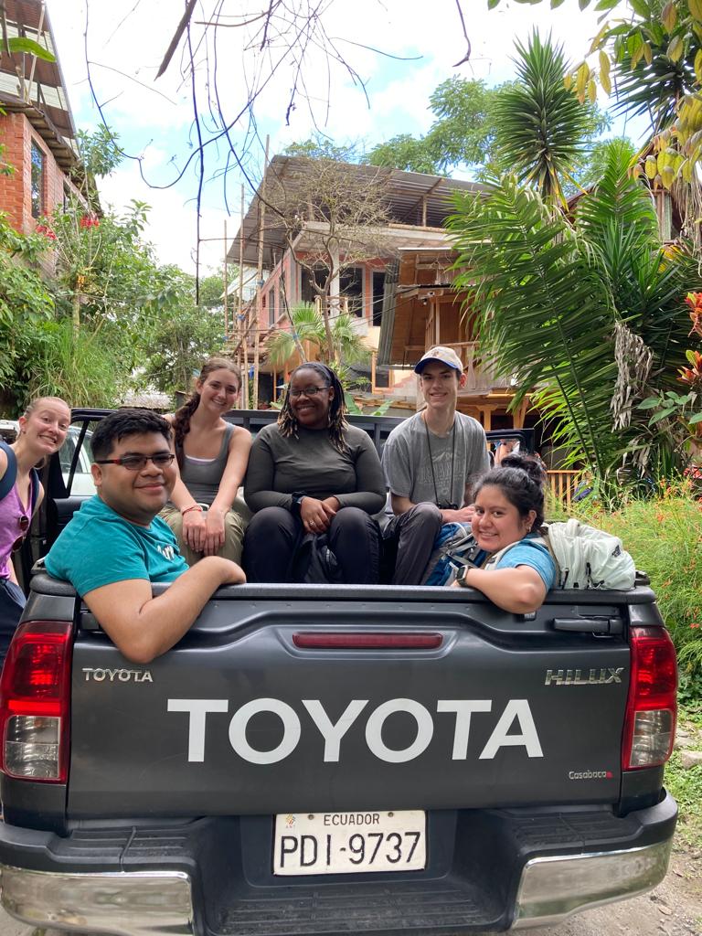 A group of young people riding in the open bed of a Toyota pickup truck on a village street.