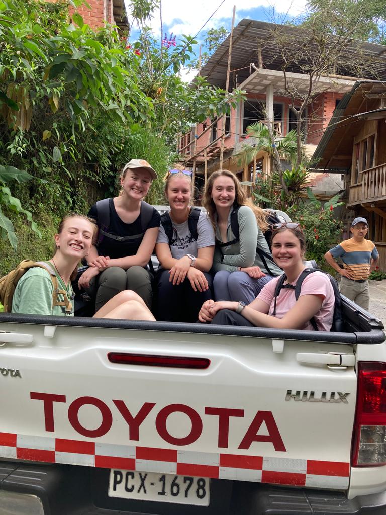 Smiling young people sitting together in the bed of a red-and-white Toyota truck with buildings behind them.