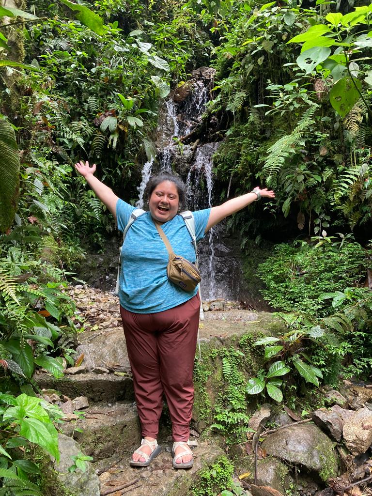 A smiling woman standing with arms outstretched in front of jungle greenery.