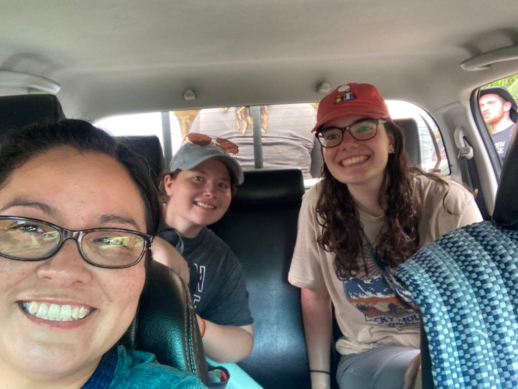 A selfie of three smiling young women sitting inside a vehicle.