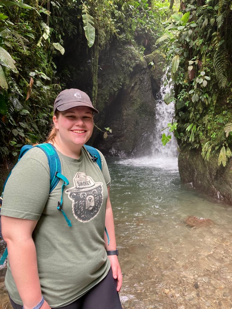 A smiling person standing beside a stream in a forested canyon.