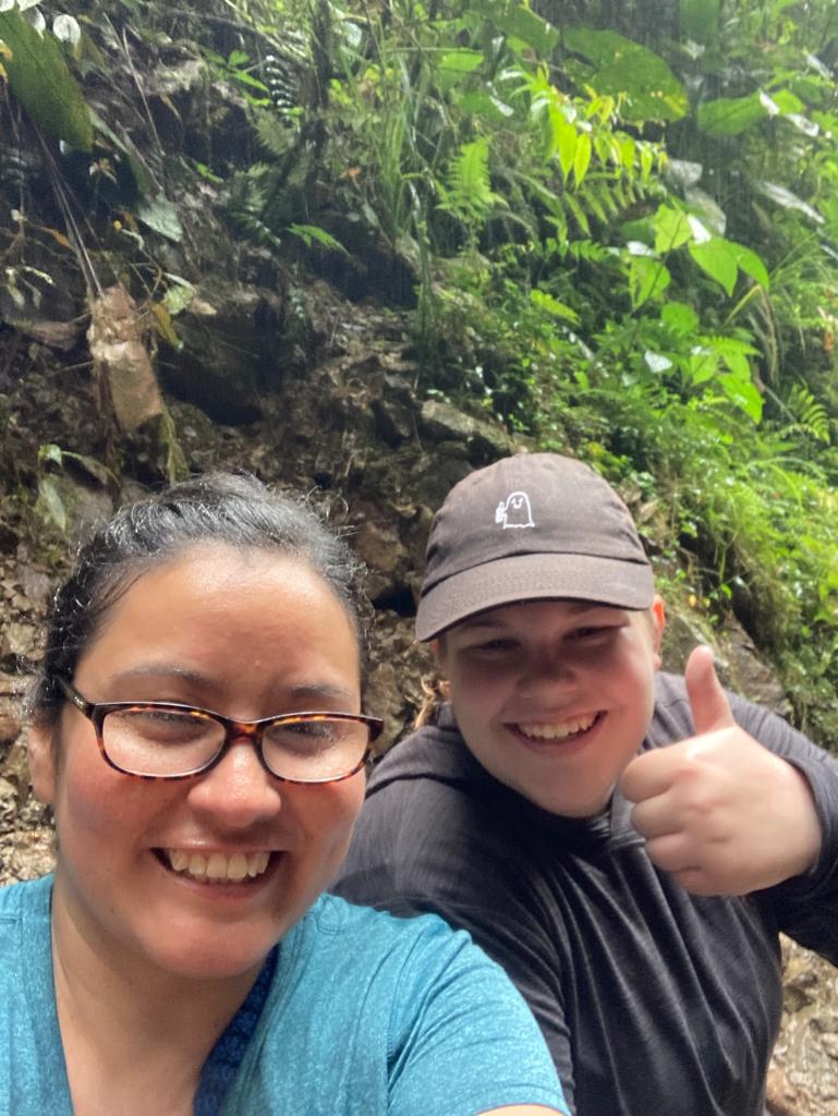 Two people taking a selfie together in the rainforest, one giving a thumbs up.