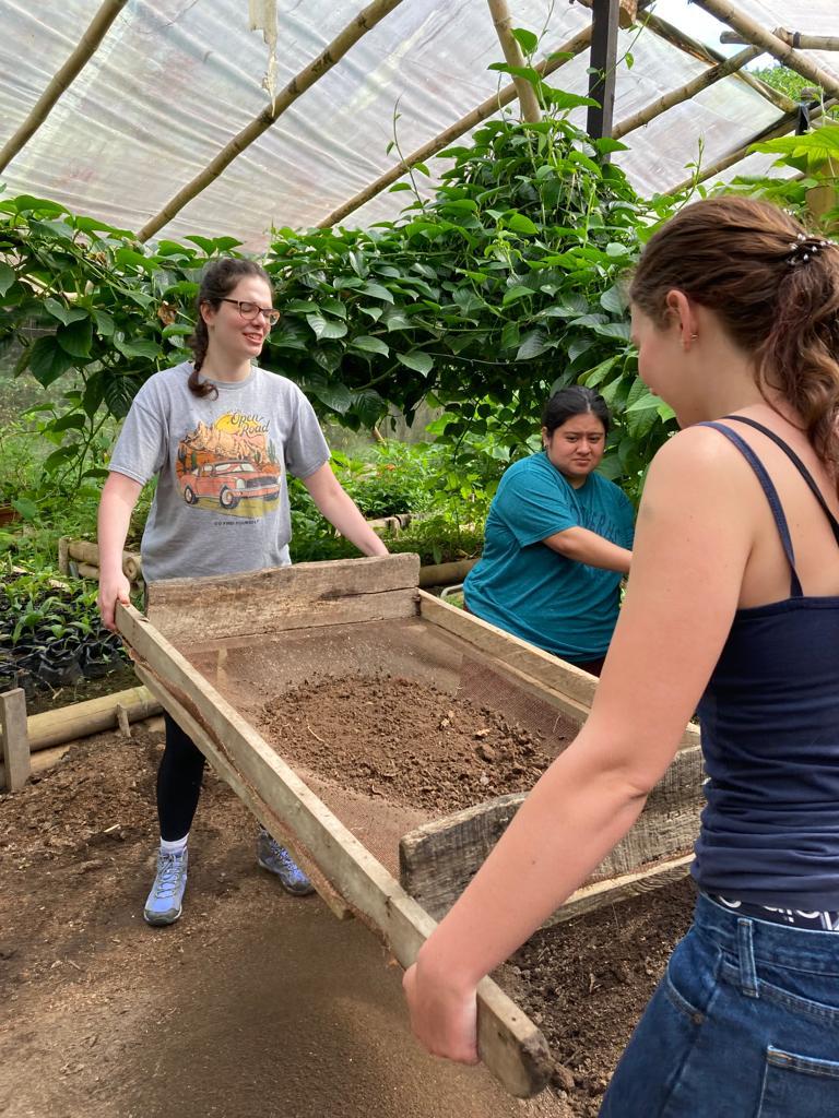 Two people holding a wooden sifting tray filled with soil in a greenhouse.