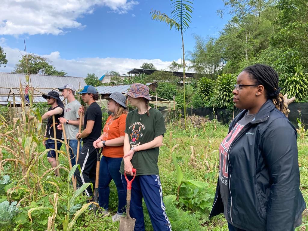A line of students working in a garden while an instructor speaks to them.