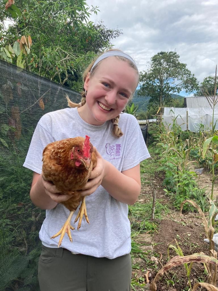 A smiling person holding a brown chicken in a garden.