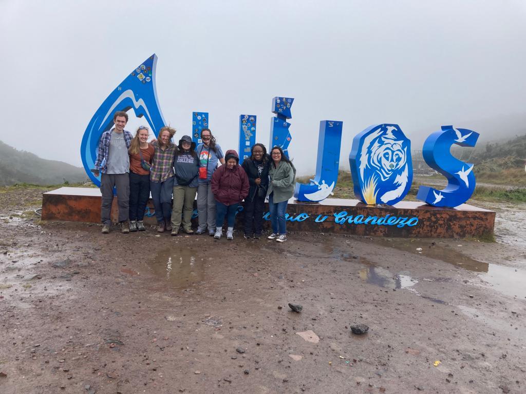 A group of people posing in front of large blue letters standing on wet ground in a mountainous landscape.