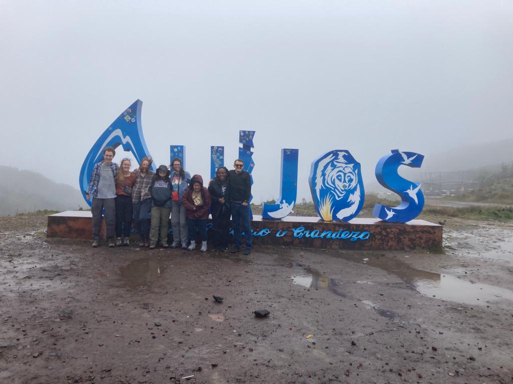 A group of people posing in front of large blue letters standing on wet ground in a mountainous landscape.