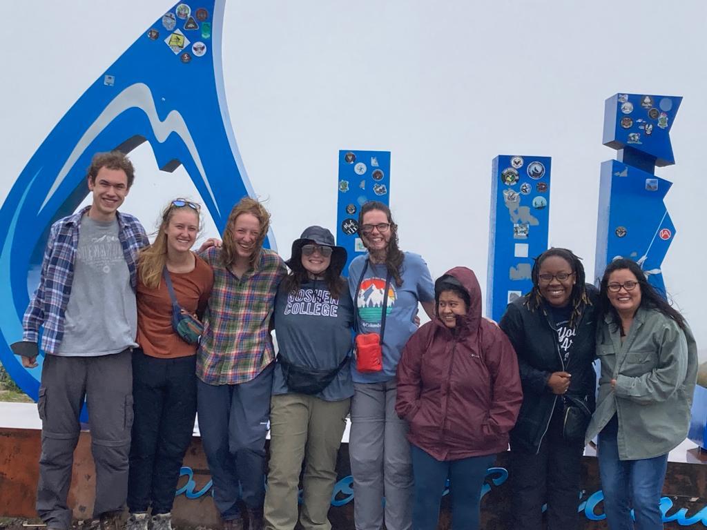 A group of people posing in front of large blue letters standing on wet ground in a mountainous landscape.