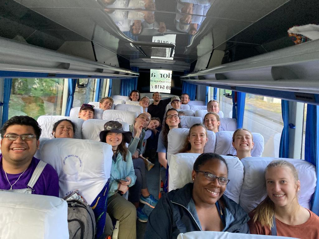 A group of smiling young adults seated inside a bus, looking toward the camera during a road trip.