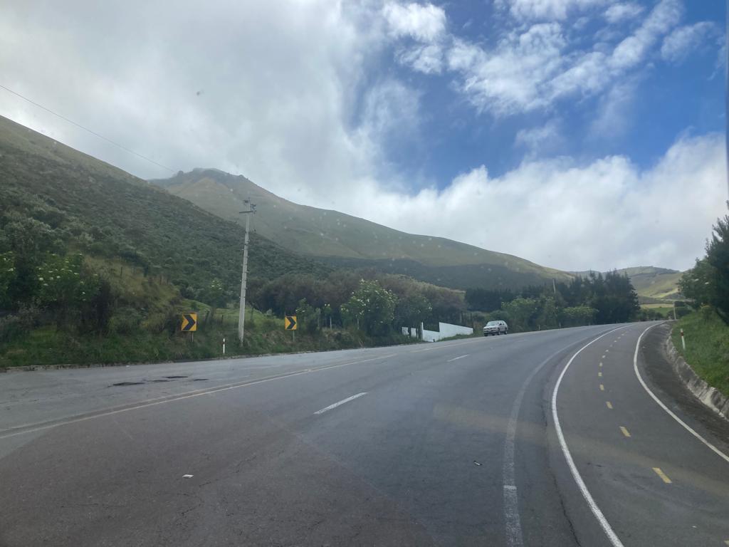 A curved mountain road with hills in the background, road signs, and a partly cloudy sky.