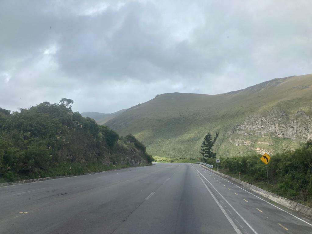 A curved mountain road with hills in the background, road signs, and a partly cloudy sky.
