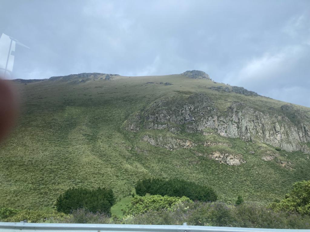 A steep green mountainside with exposed rock formations and low vegetation under a pale sky.