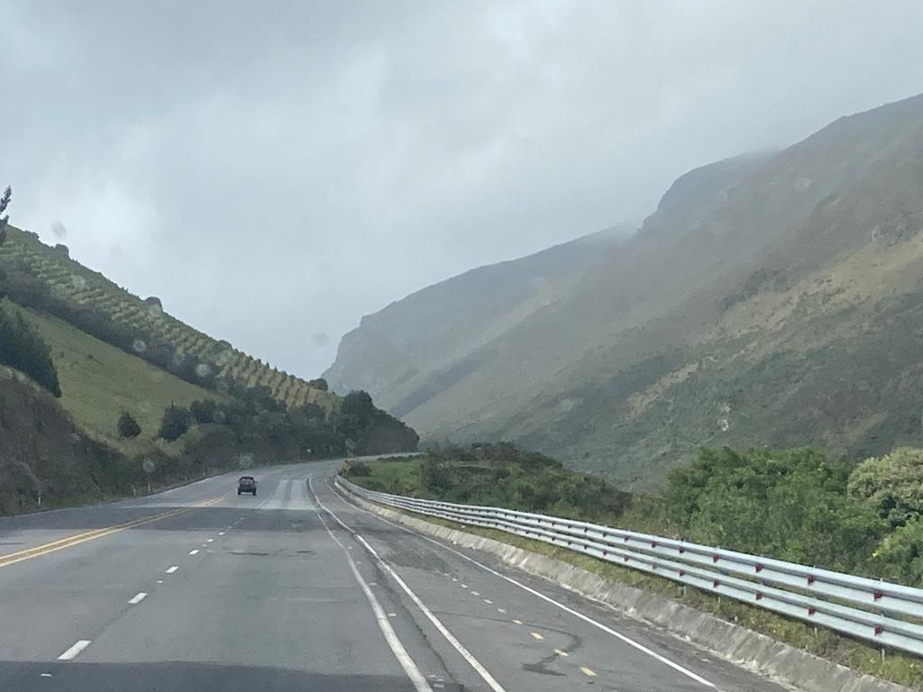 A wide mountain road flanked by hills and vegetation, with cloudy weather.