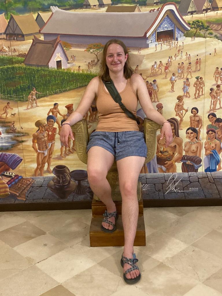 A woman posing seated in front of a mural depicting indigenous life.