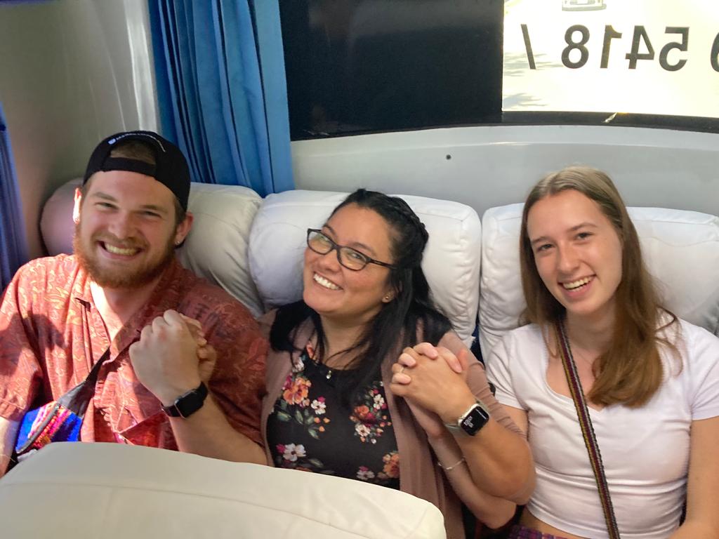 Three students smile together in the reclined seats of a tour bus.