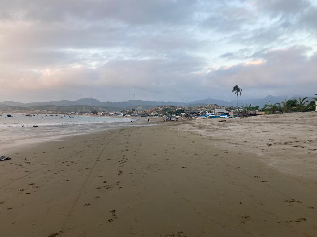 A wide sandy beach at dusk with a town and hills in the distance.