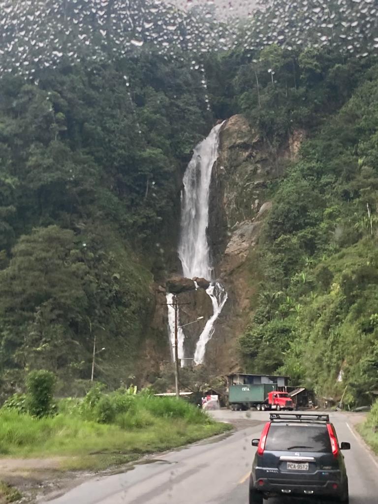 A tall waterfall beside a mountain road with cars driving past.
