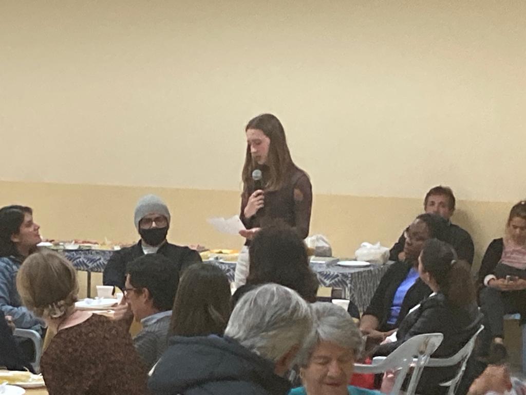 A woman addressing a seated audience at an indoor meeting.
