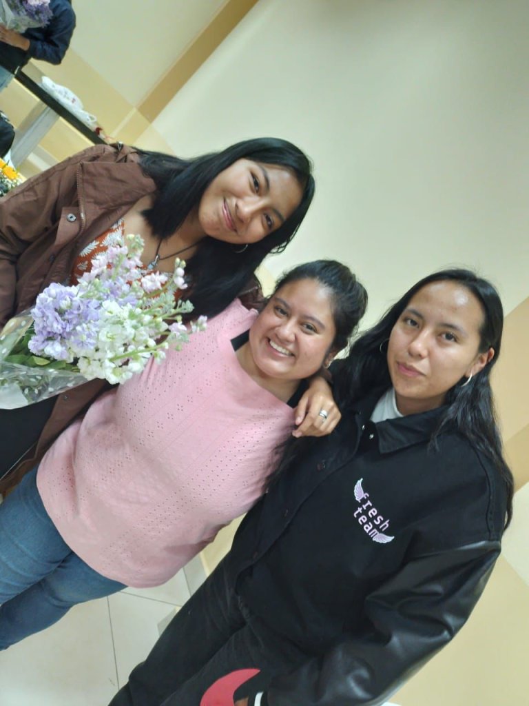 Three women posing together, one holding flowers.
