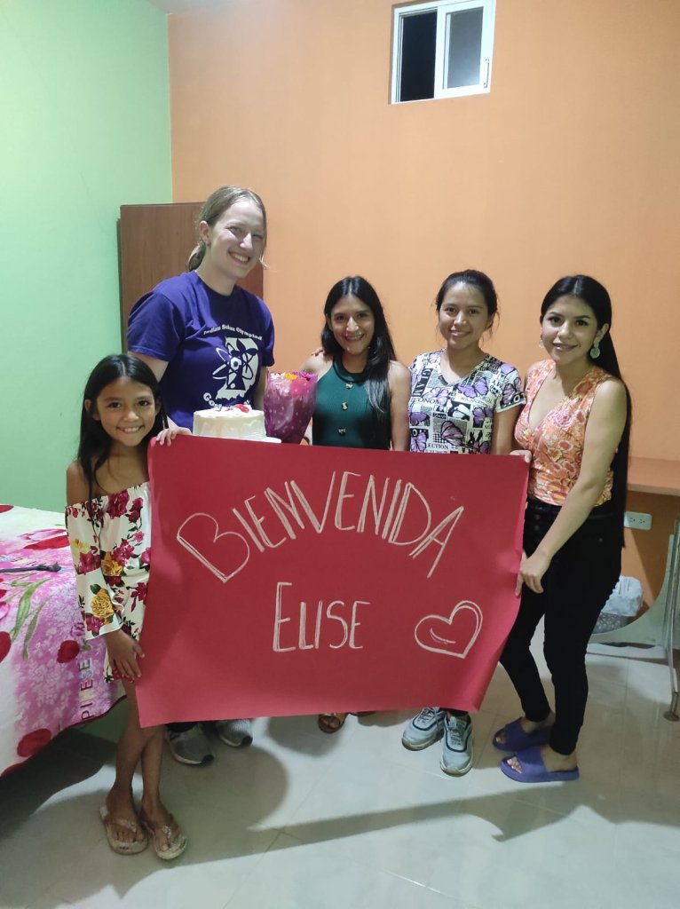 A student holding a "Bienvenida" welcome banner with a host family.

