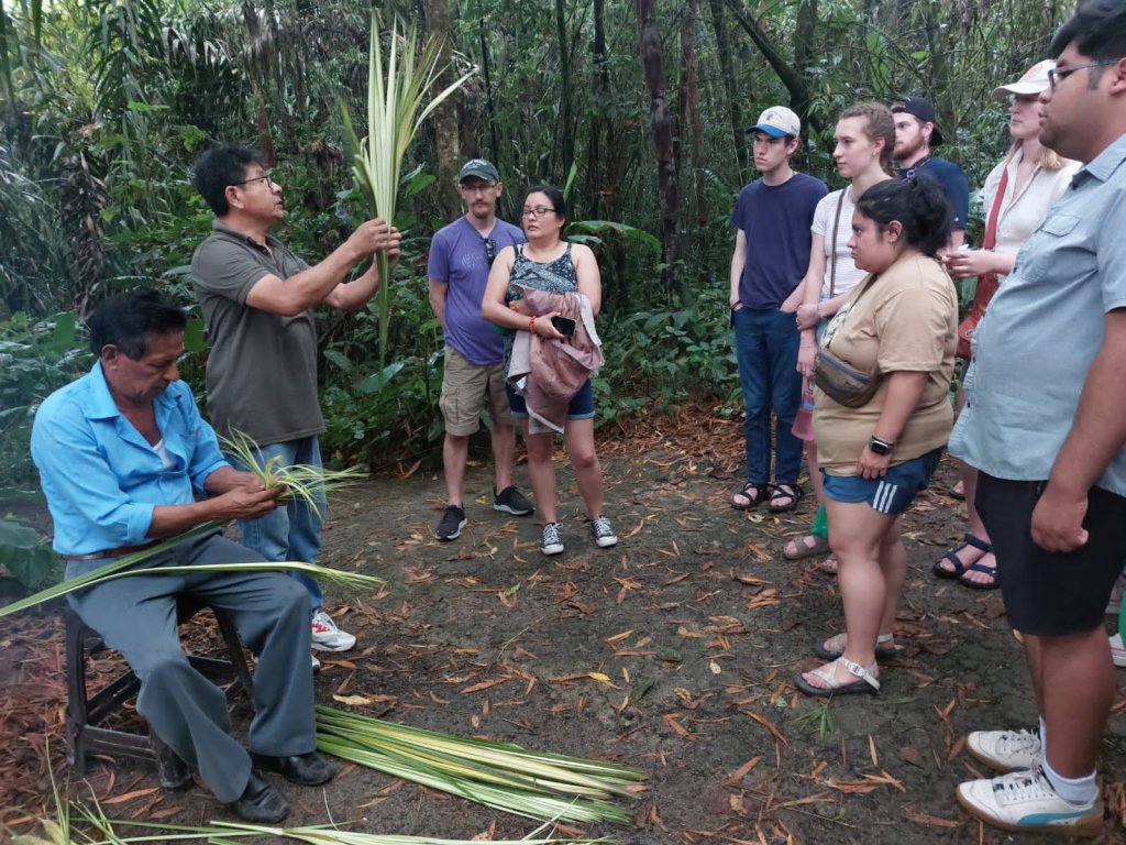 A group watching a seated man prepare palm fronds on the ground.
