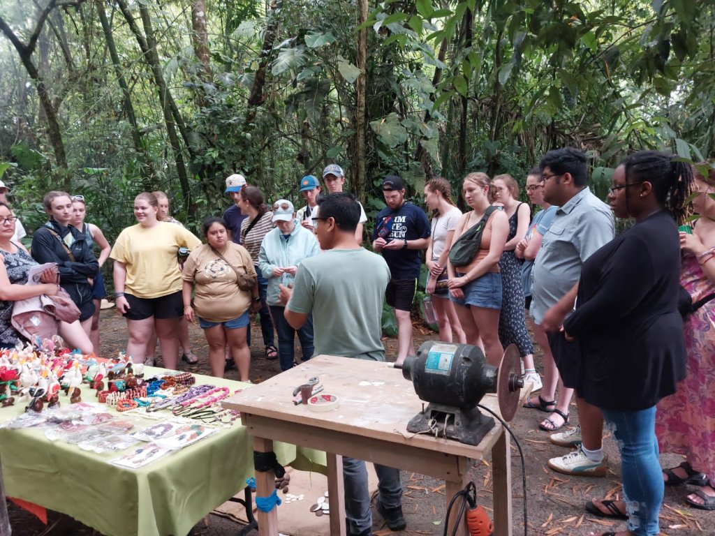 Students browsing a table of handmade crafts in the forest.
