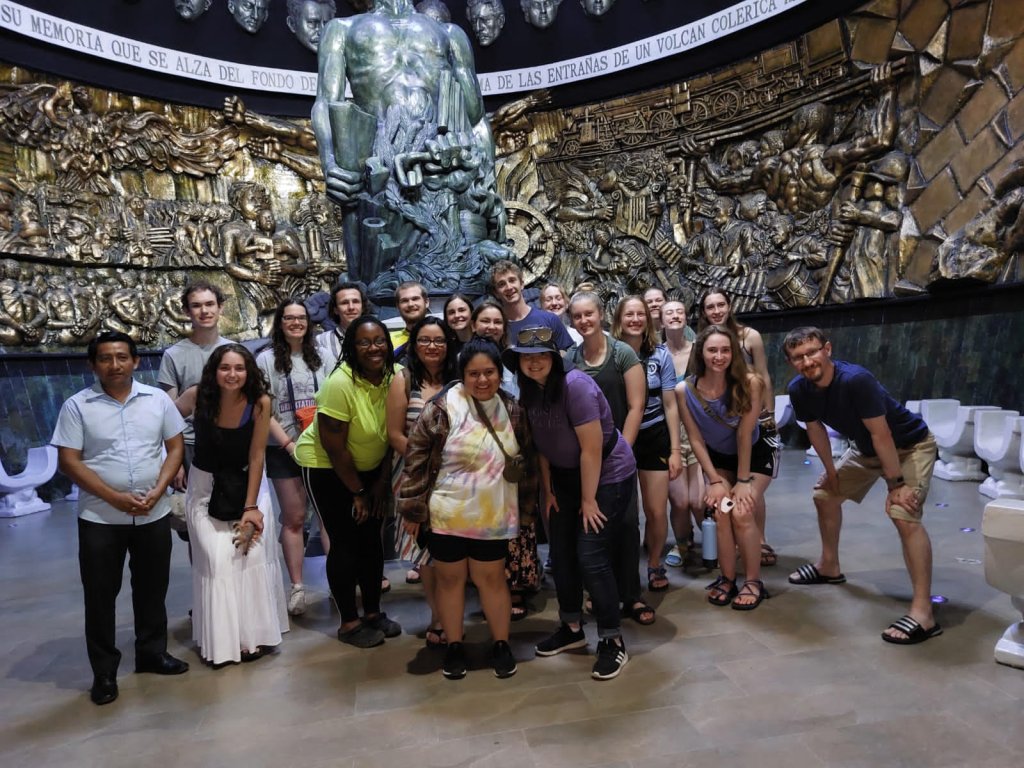 A whole-group photo in front of a large bronze relief mural.
