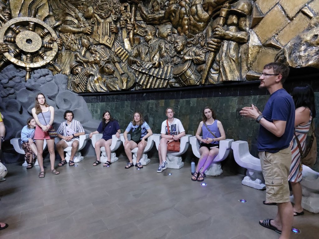A group of students sitting along a wall as a guide speaks.
