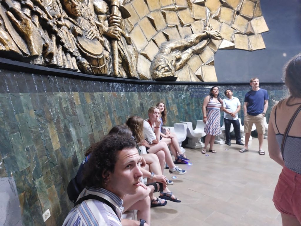 Students seated near an ornate gilded wall in a gallery.
