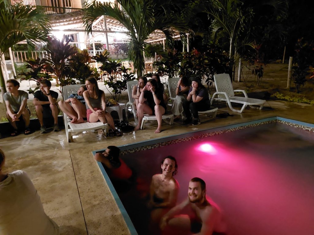 Students relaxing in a pink-lit swimming pool at night.
