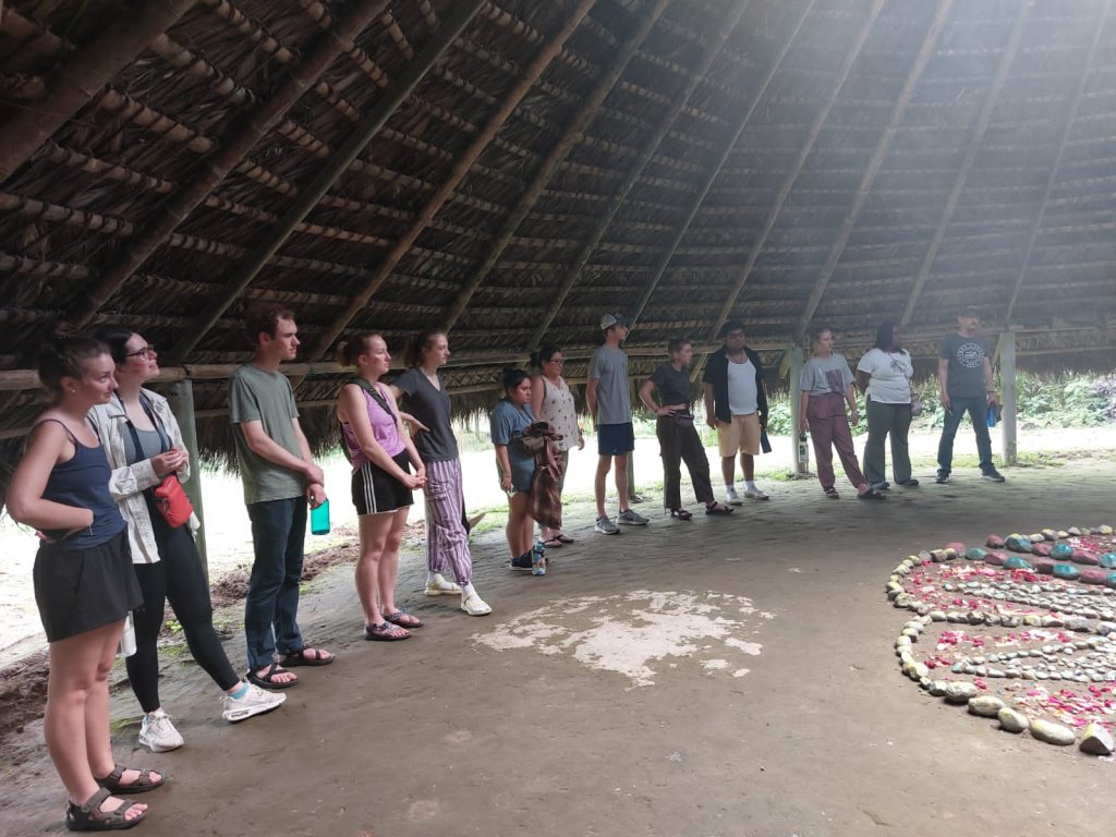 Students standing around a colorful ground mandala in a thatched hall.
