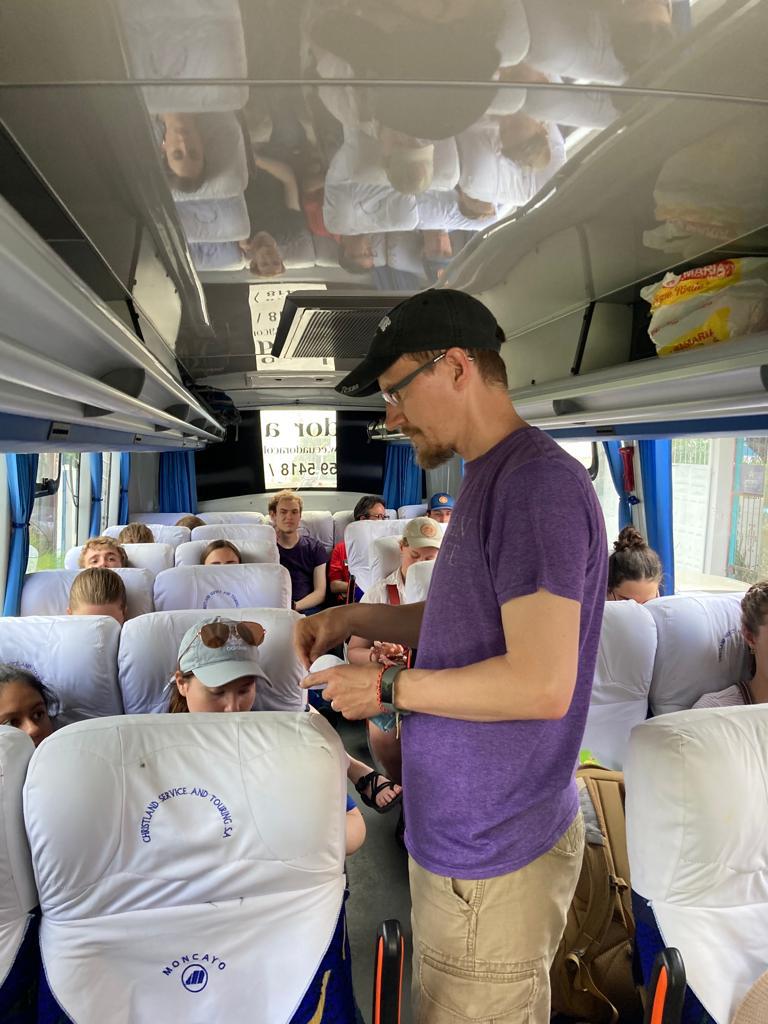  young man in a purple shirt and black cap stands in the aisle of a tour bus while other travelers sit in the seats around him.