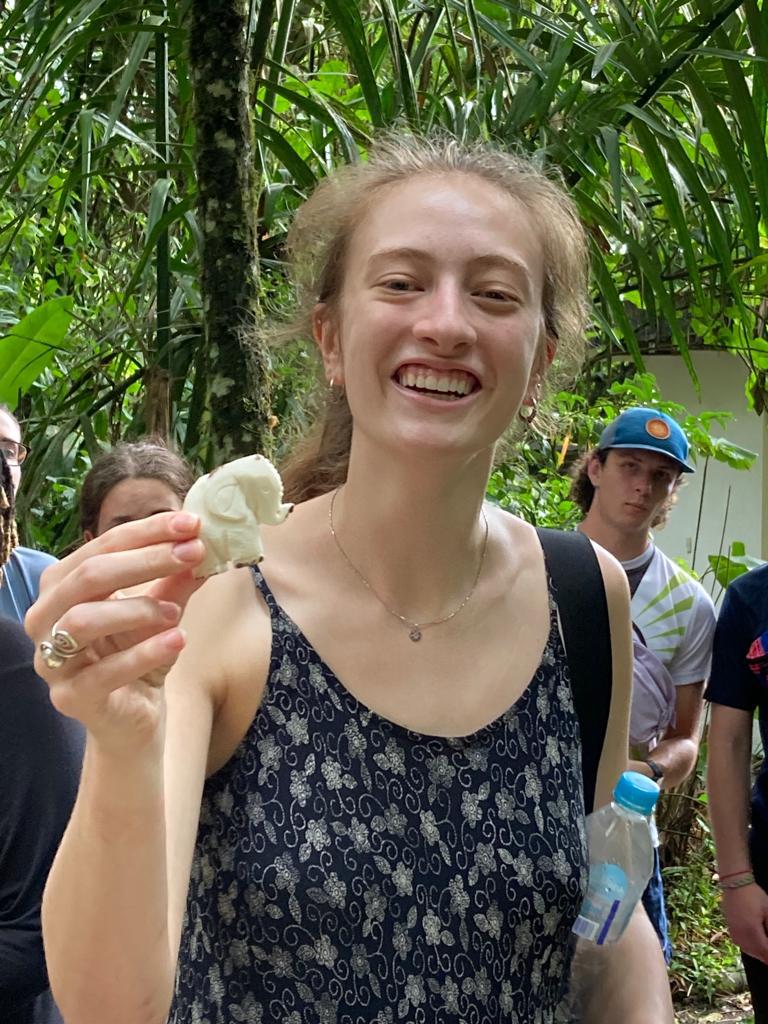 A smiling woman holding up a small woven item outdoors.
