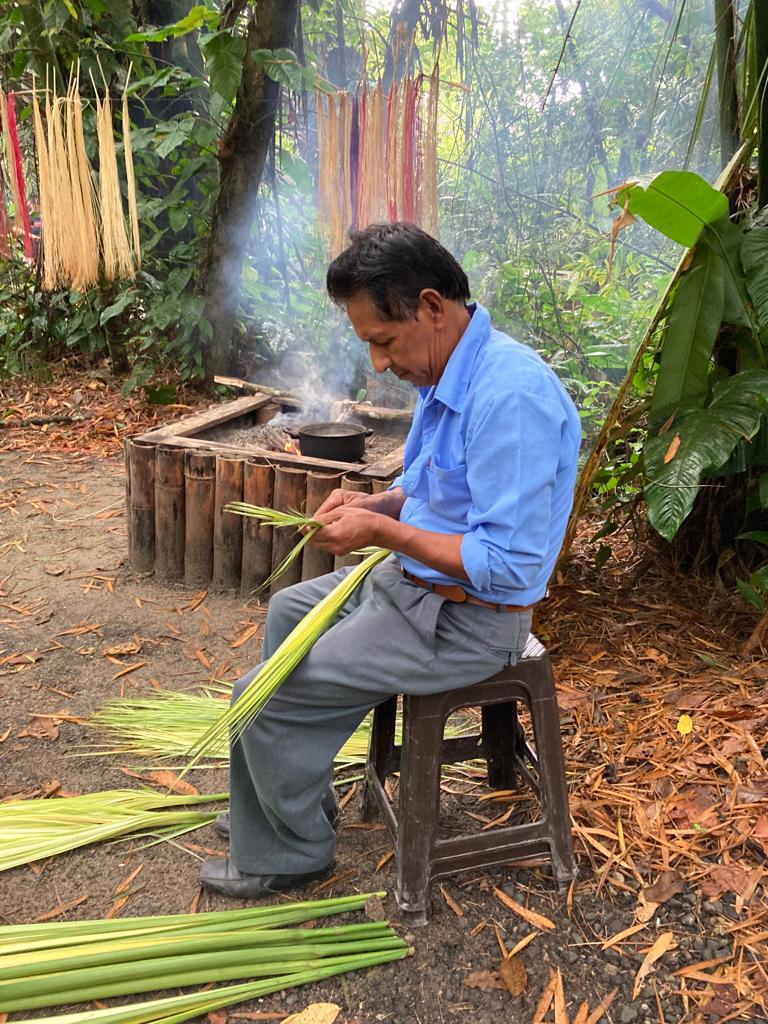 A man seated stripping palm leaves beside a smoking fire.
