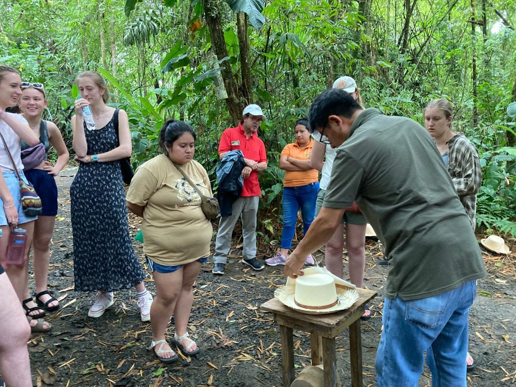 A guide shaping a hat on a wooden stand as students watch.
