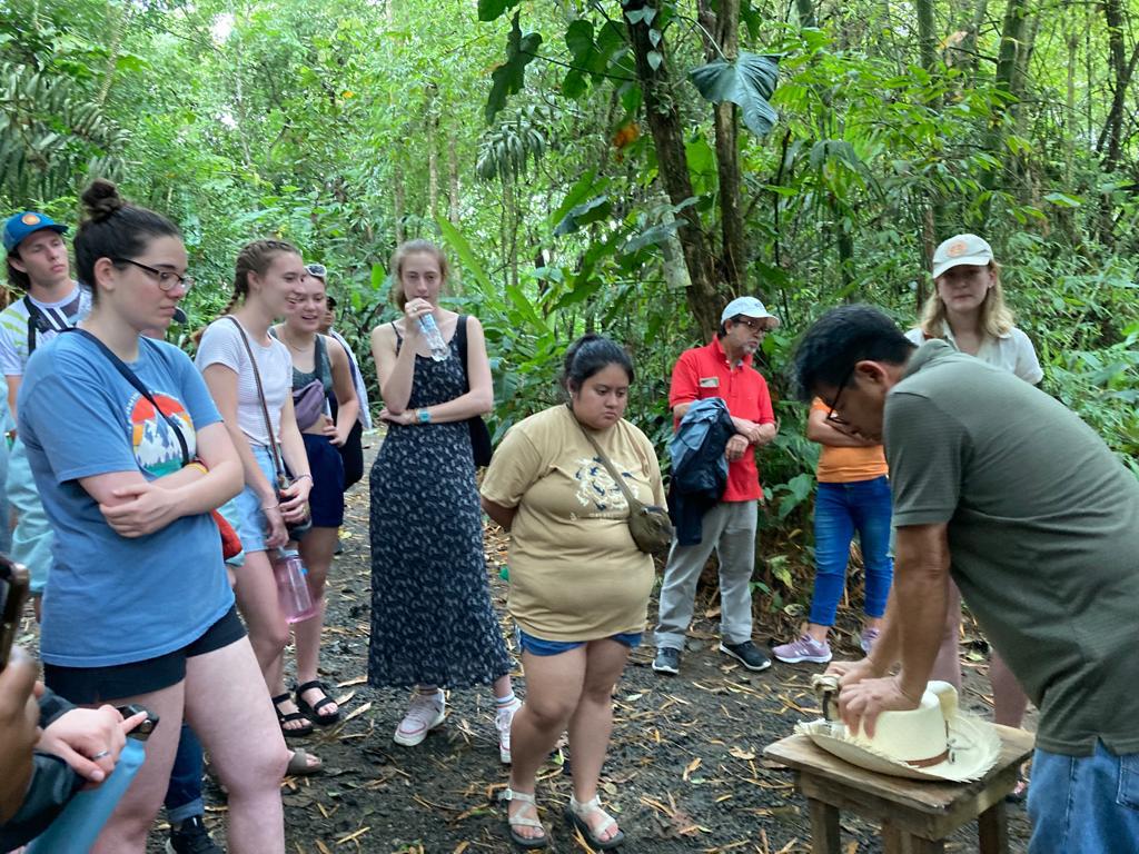 A group watching a hat-shaping demonstration on a wooden stump.
