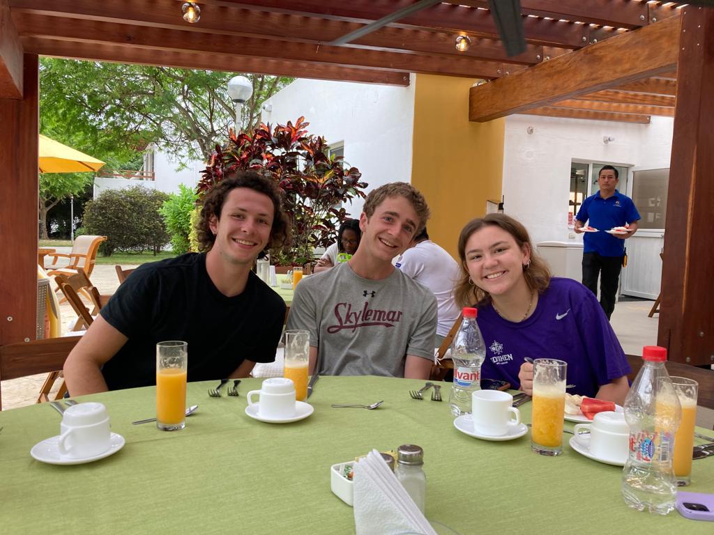 Three students smiling at an outdoor breakfast table.