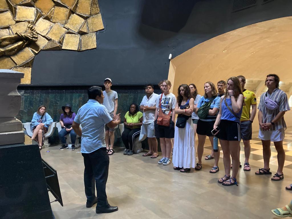 A group of students listening to a guide inside a dim gallery.
