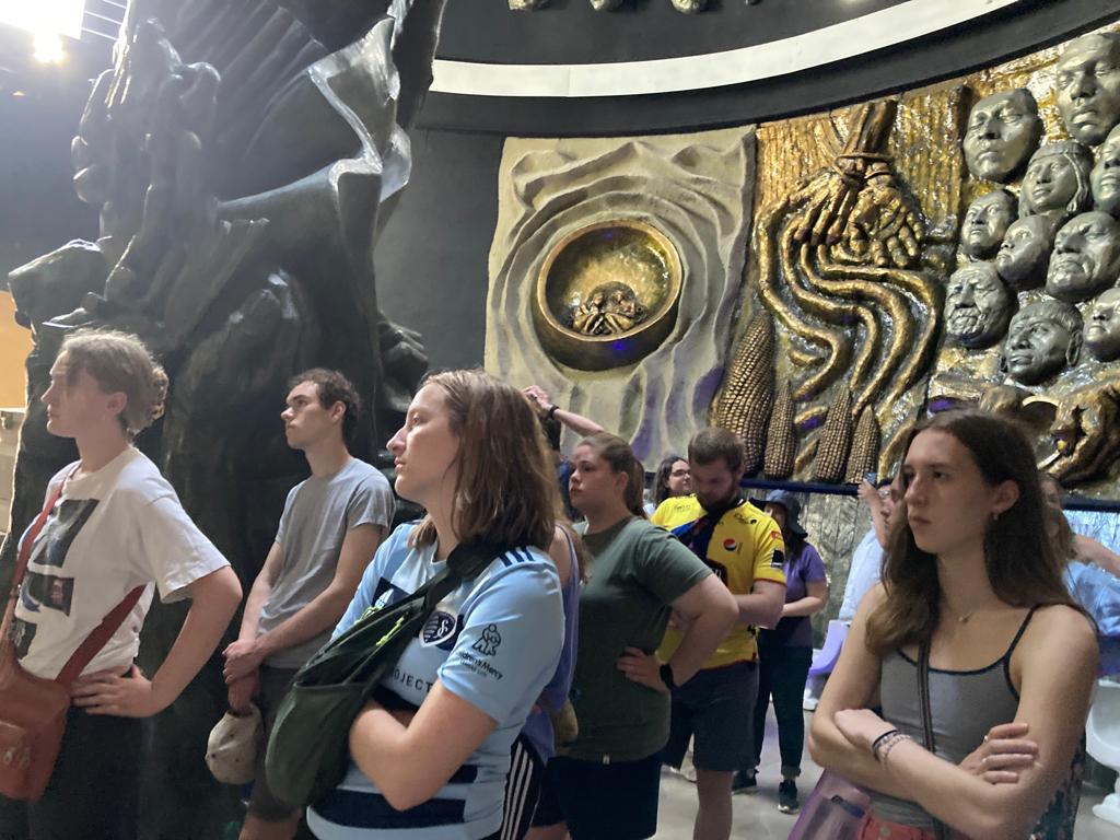 Students viewing a large bronze relief sculpture in a museum.
