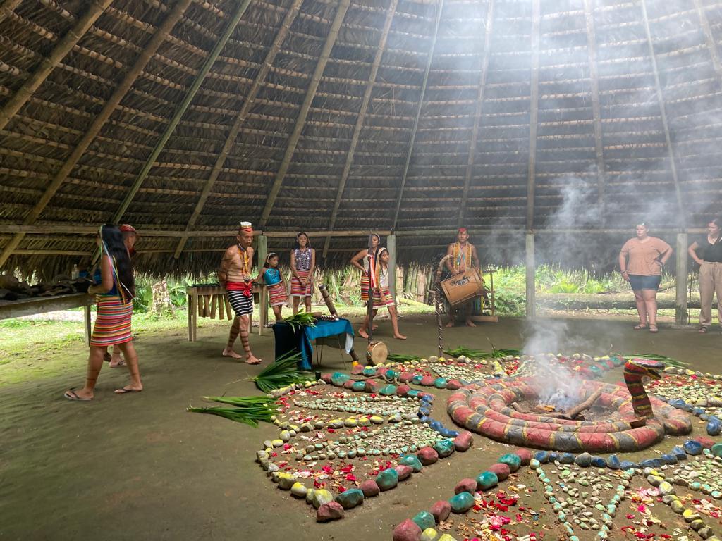 Performers around a ceremonial mandala with smoke rising in a hut.
