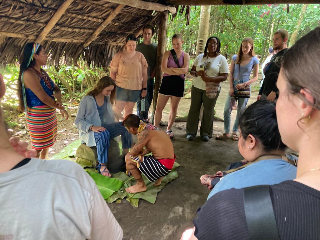 Students watching an indigenous cultural demonstration in a thatched hut.
