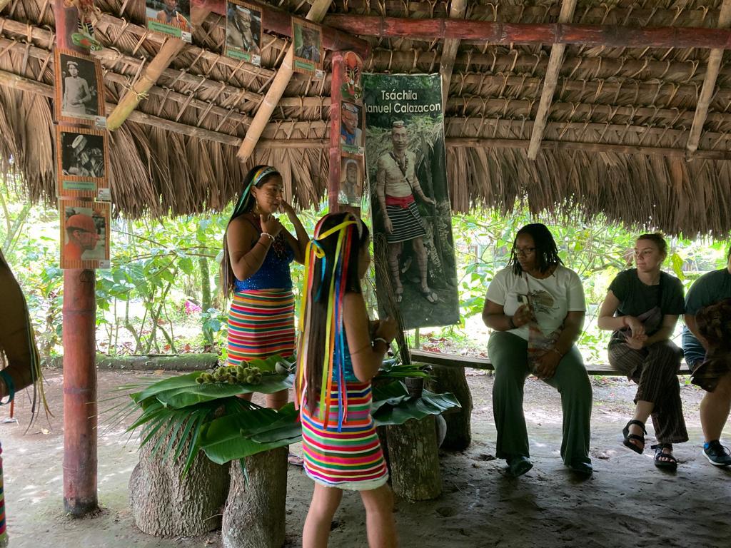 A woman in traditional dress presenting to students under a thatched roof.
