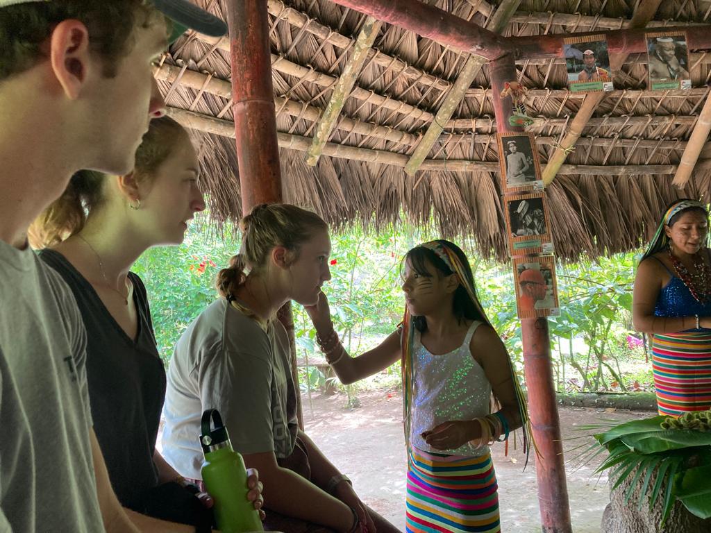 A woman in traditional dress interacting with students in a thatched hut.
