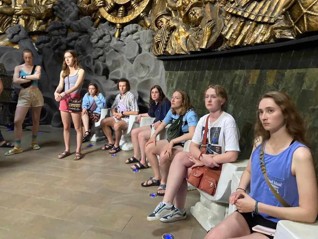 Students seated listening in a museum room with golden reliefs.
