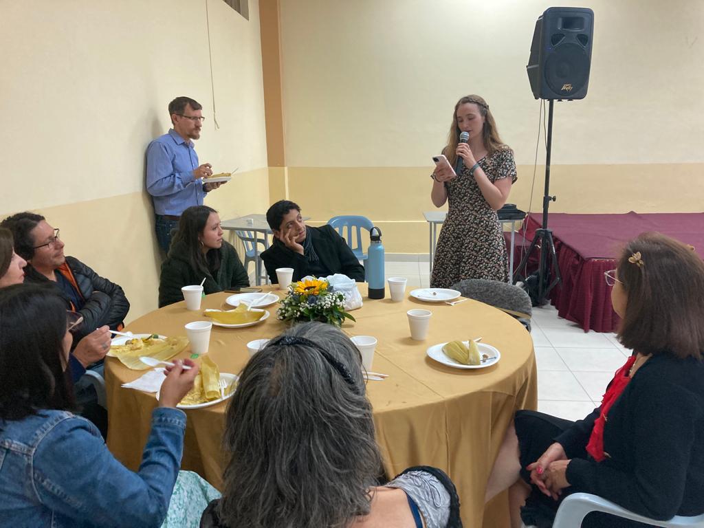 A woman speaking to guests seated at a table in a hall.
