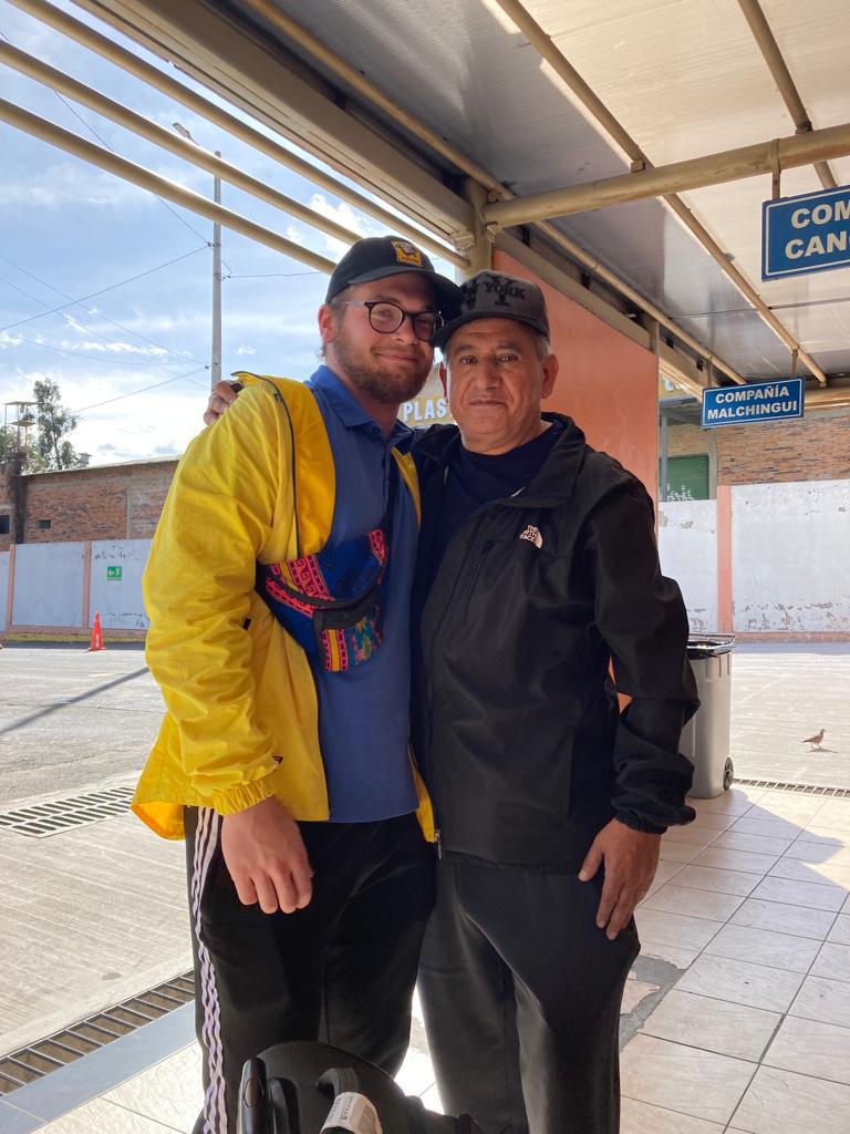 Two men posing together at an outdoor bus station.
